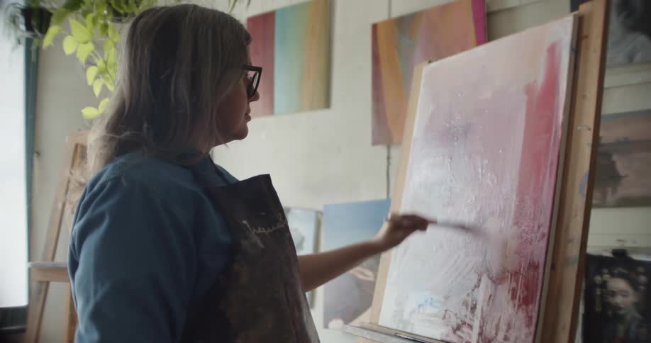 A female painter in an apron uses a paintbrush to paint on a canvas resting on an easel indoors during the day