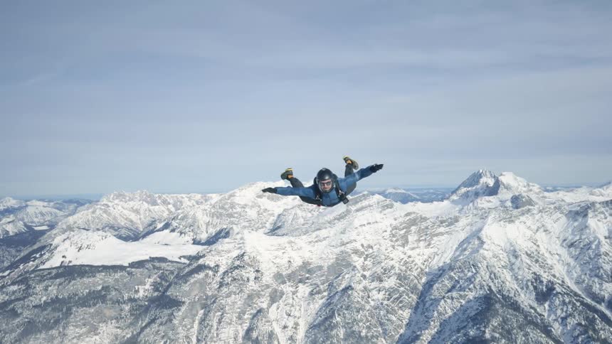 Skydiver wearing a parachute pack and black helmet is floating over snow covered mountains