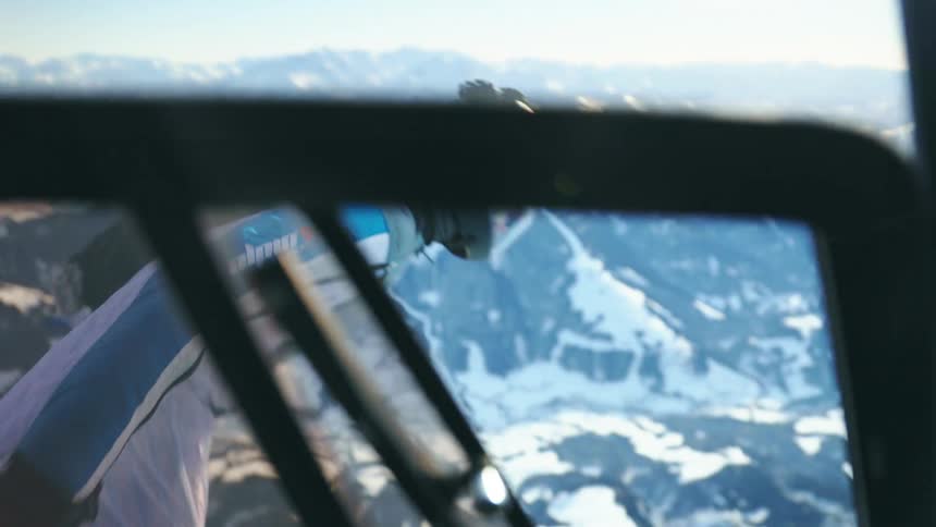 A paraglider holding onto the side of an airborne aircraft releases his grip and dives towards the snowy mountain range below