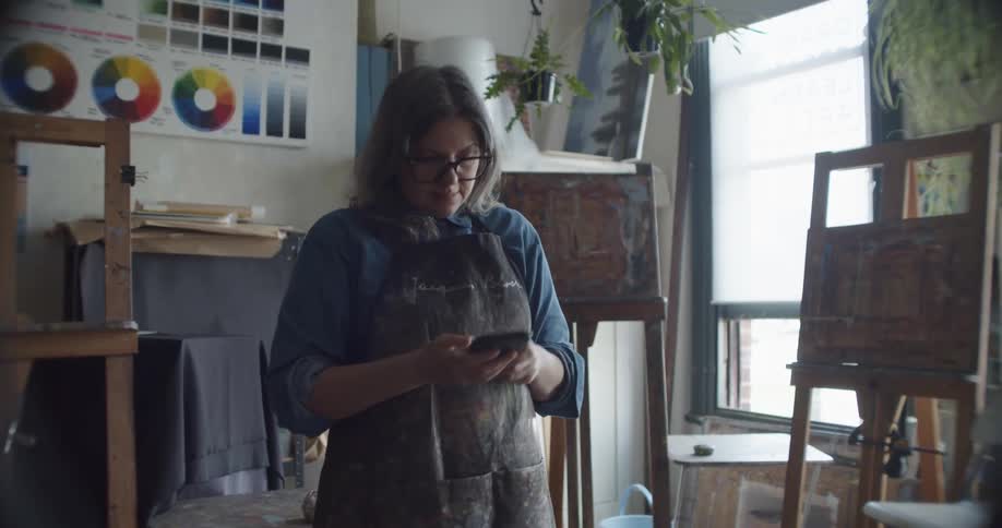 A woman pulls a smart phone out of her pocket and takes a picture of artwork on an easel inside of an art studio