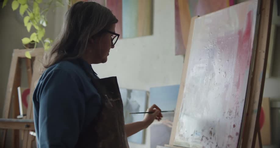 A woman in a room with glasses and an apron painting on a canvas with a paint brush