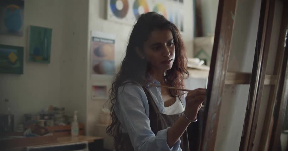 Woman wearing an apron painting with a small brush over canvas on an easel inside a studio