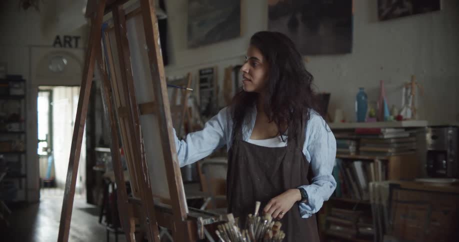 A woman looks at her artwork and picks up a paintbrush as she paints in a studio