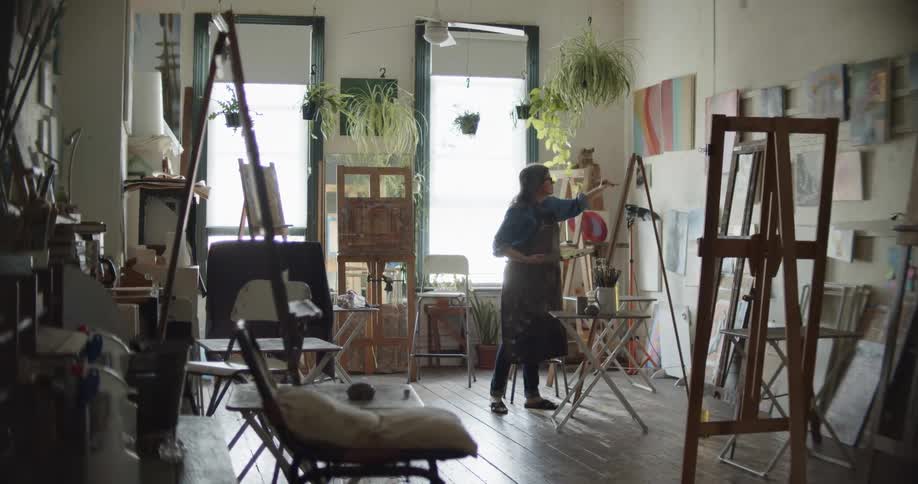 A female artist painting on a canvas in an art studio with hanging houseplants
