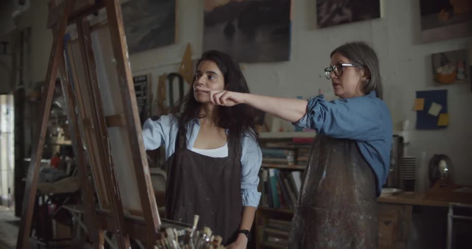 Two women in a room with one painting on a canvas with a paint brush