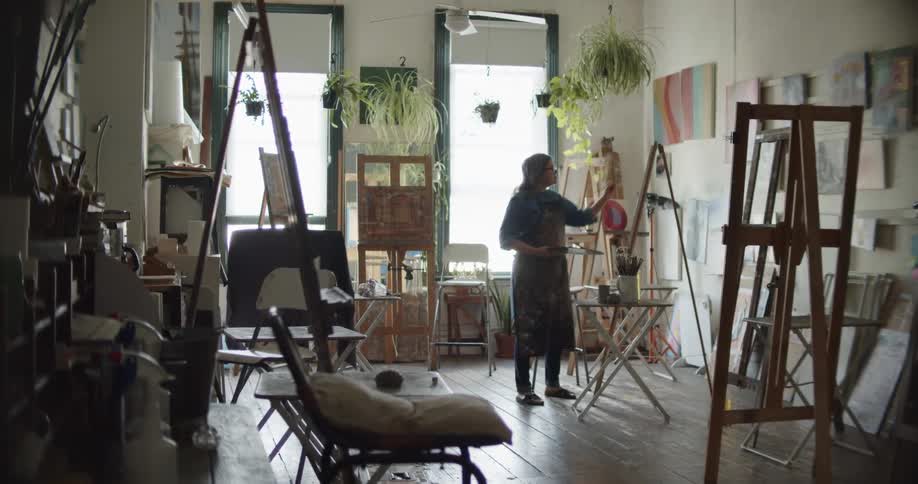 A woman holds a pallet of paint in one of her hands while she paints with a paint brush onto an easel in an art studio during the day