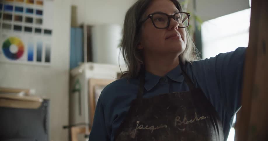A woman uses a paint brush to get paint off an art pallet and brush in onto a board on an easel