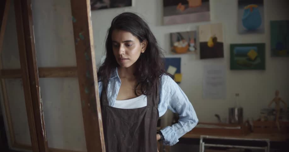 Woman wearing an apron painting with a small brush over canvas on an easel inside a studio