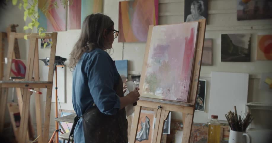 A woman looks at her painting and then runs her paint brush over it