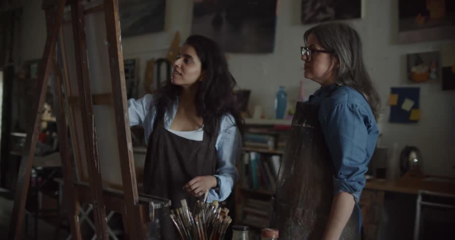 A woman teaches another woman how to paint inside of an art studio during the day