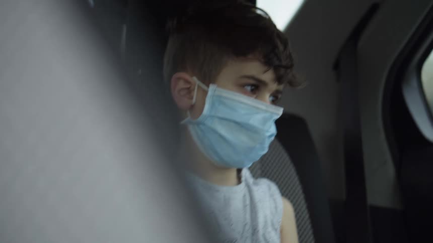 A young boy sitting in a car pulls a mask up over his mouth and nose