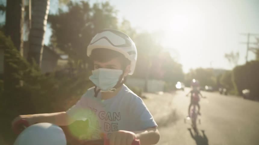A boy with a helmet and mask riding a bike with training wheels with the sky and clouds in the background