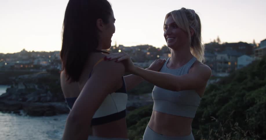 A pair of women stretch while holding each others shoulder for balance on a cliff overlooking the ocean in the evening