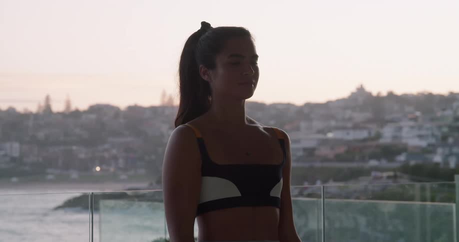A young woman lifts dumbbells while standing outdoors on a balcony overlooking the coast of a city during the day