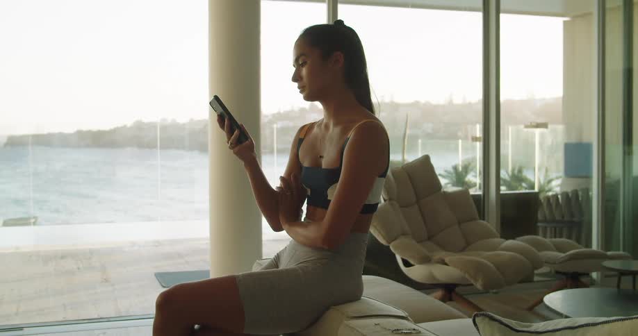 A woman sitting down in a room looking at a smart phone with windows in the background