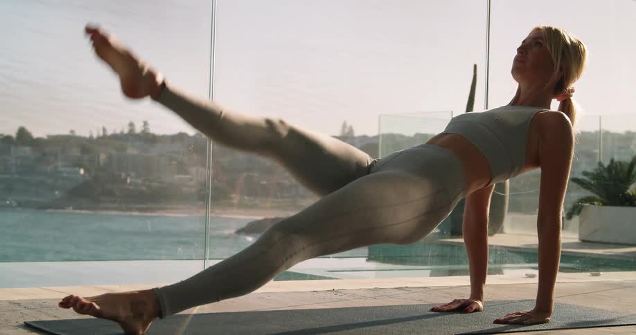 A woman does a yoga stretch and leg lifts on a mat next to large windows with an ocean view