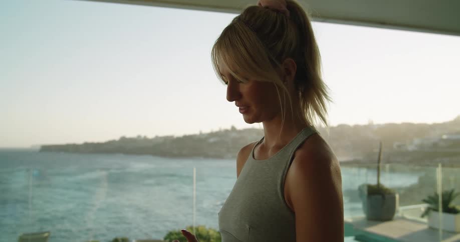 Young fit woman wearing sports clothes using her smartphone near the pool at home close to the ocean