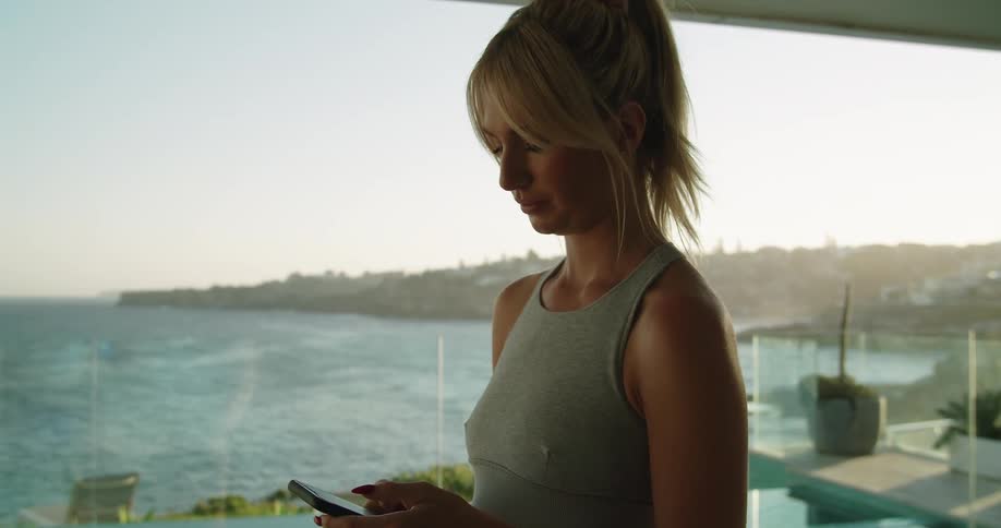 A woman in a balcony looking at a smart phone with the sky and clouds in the background