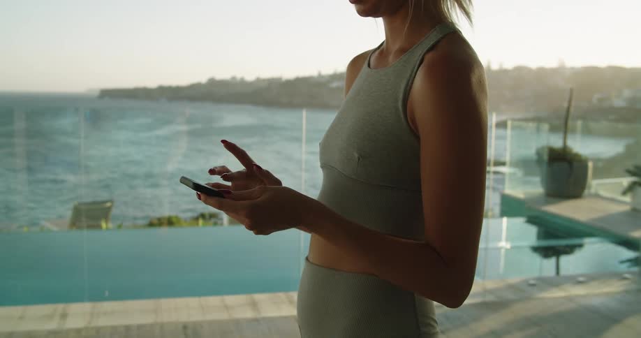 Young woman wearing sports clothes checking her smartphone at home near the ocean at sunset
