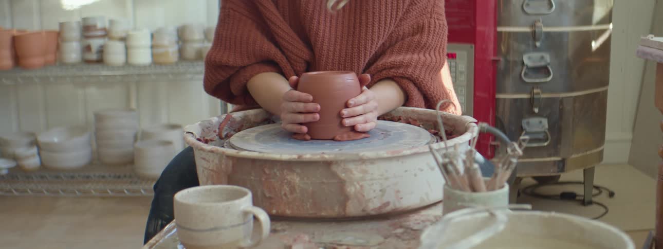 Young woman wearing red sweater pics up a freshly turned clay vase from the turning wheel