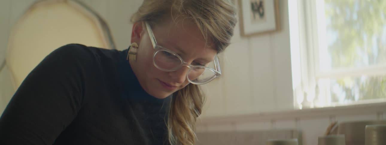 A woman wearing glasses focuses on her project while she sits in a pottery crafting studio