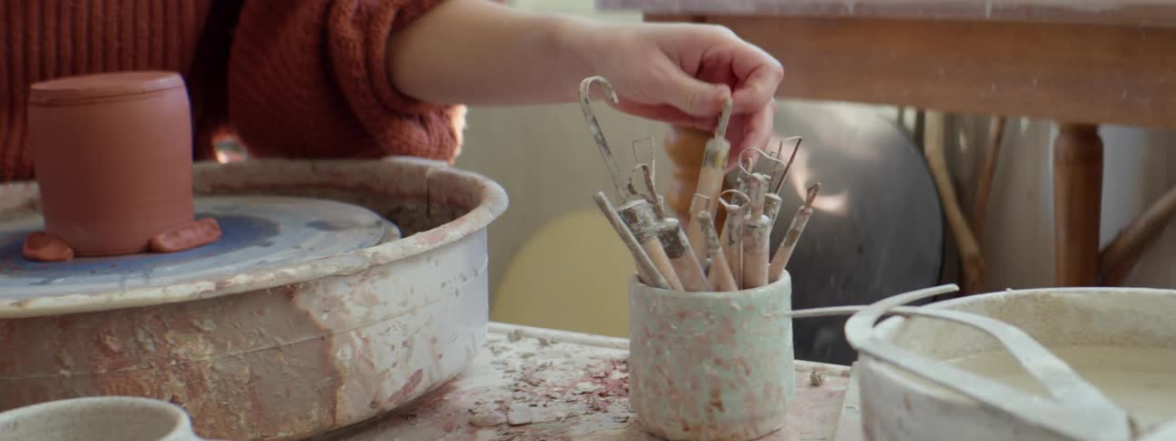 A woman picking up a tool while working on a pottery wheel