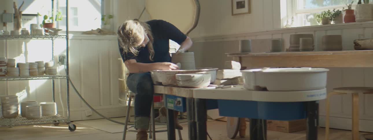 A woman sitting in a pottery wheel shaping a vase inside a studio