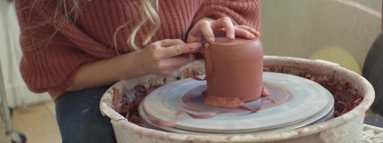 A sculptor uses a tool to shape clay on a spinning potters wheel indoors during the day