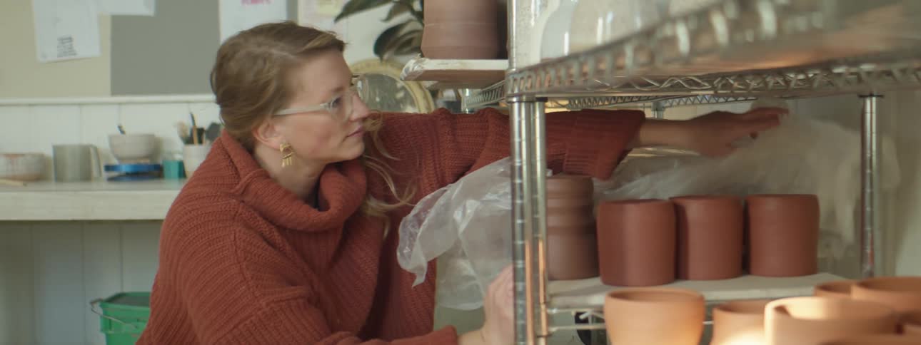 Woman wearing red sweater uncovers fresh pottery on a shelf in her studio