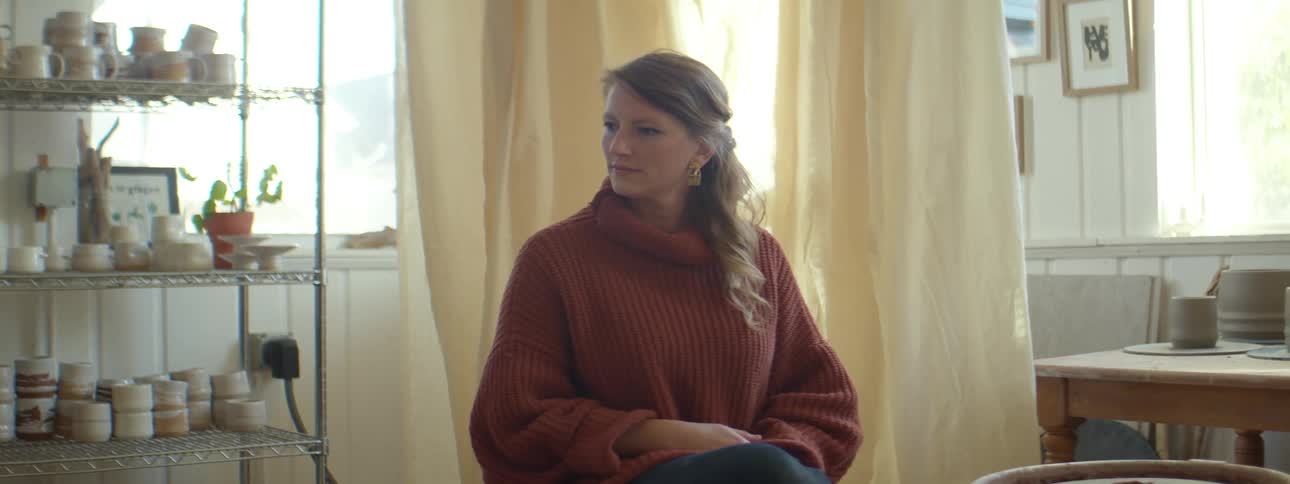 A woman with a sweater sitting in a room with a window and shelves in the background
