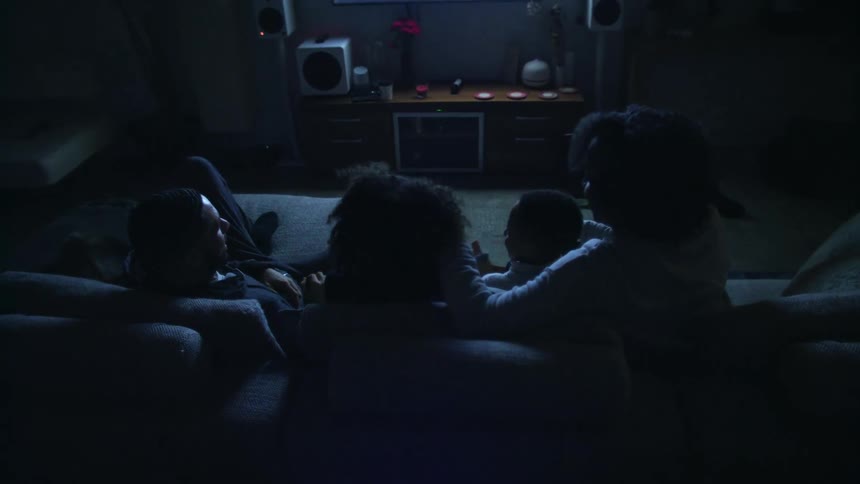 A family sits down together on the sofa of a dark living room illuminated by a large television