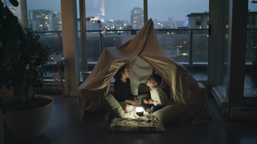 A young brother and sister sit in a fort in their home and laugh as they play hand games together