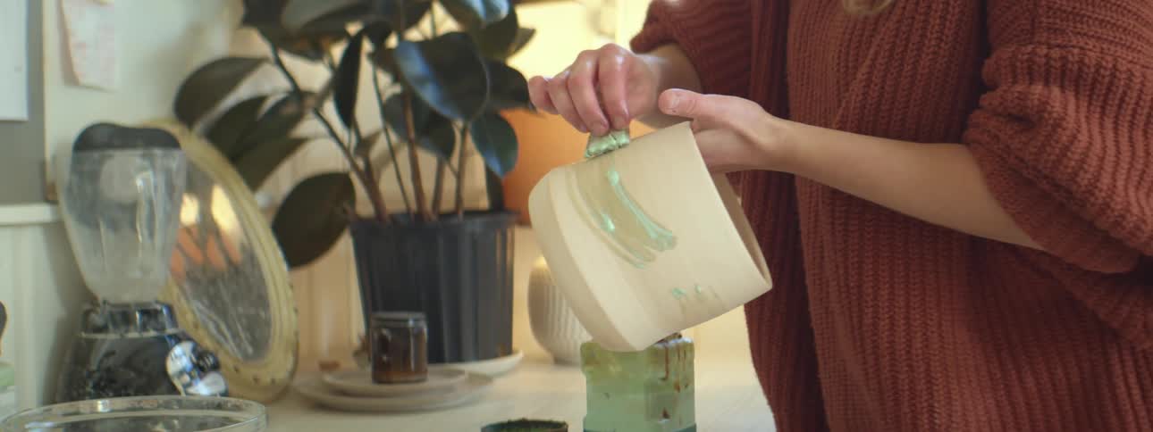 A woman wearing a baggy red sweater carefully applies a liquid coating onto a ceramic pot