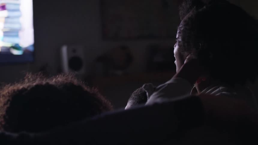 A woman watching television in a dark room with her children looks down to gaze at the child leaning against her