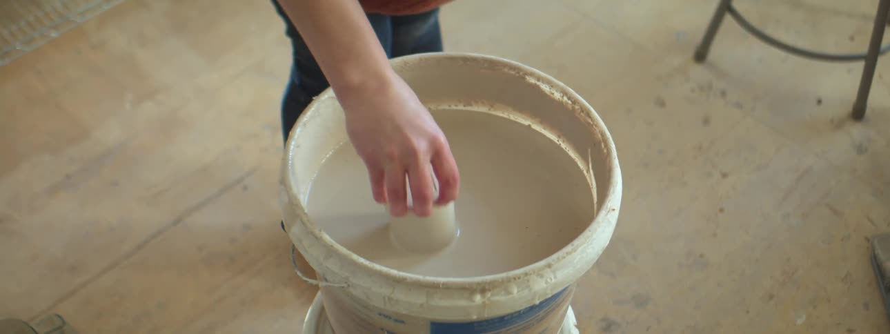 A woman dips a ceramic vase in a bucket of white paint