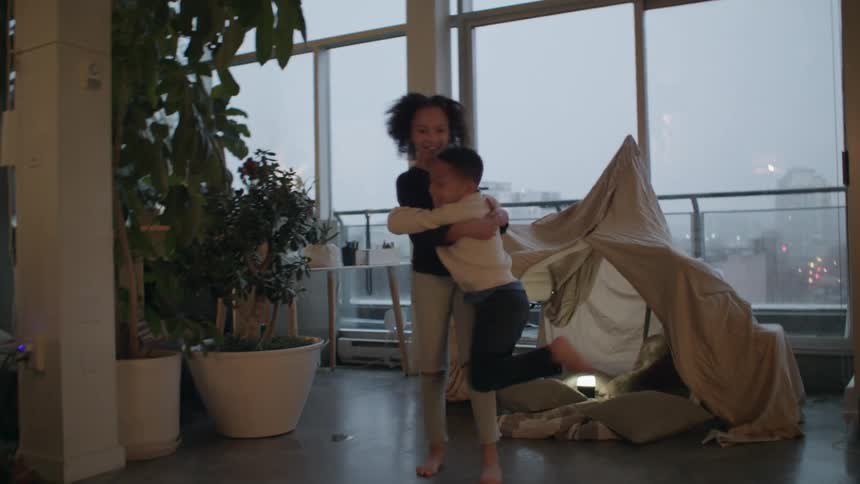 A boy running and jumping as a girl picks him up and swings him around with plants and windows in the background