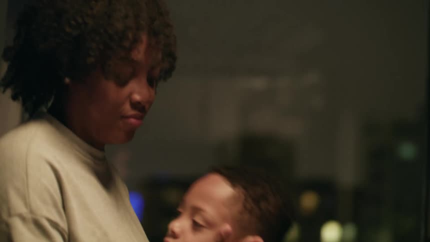 Mother kisses and caresses her son standing next to a window at home at night