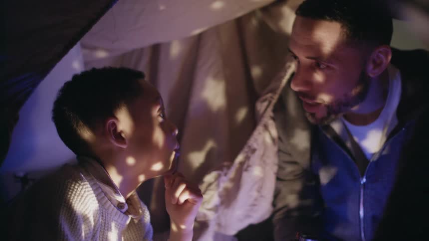A father and two children playing in a blanket fort in the dark