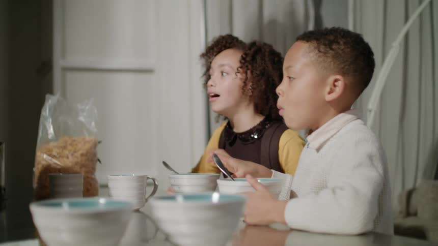 Young siblings sitting on the kitchen table eating breakfast and talking with their parents