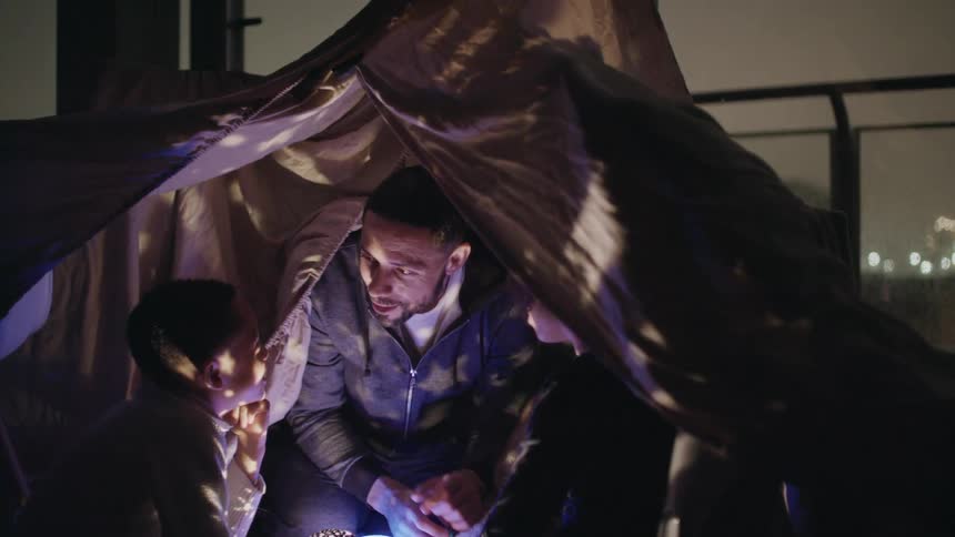 A man telling two children a story under a tent in a building with large windows overlooking a city