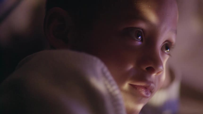 A young boy leans against his hand and talks as lights reflect on his face