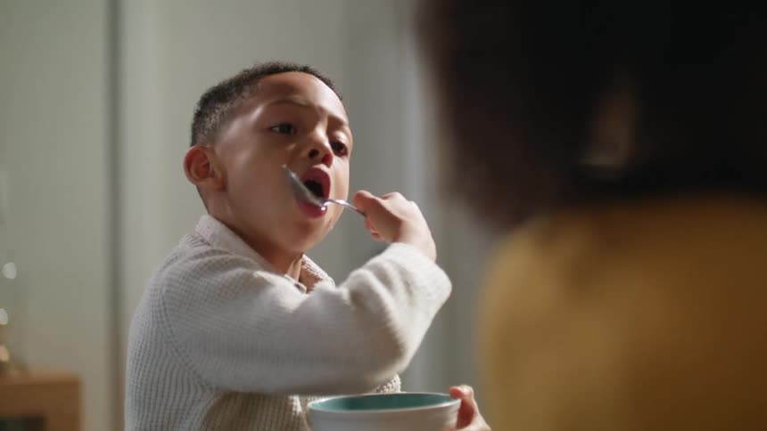 A boy eats cereal from a bowl while sitting at a dining table during the day