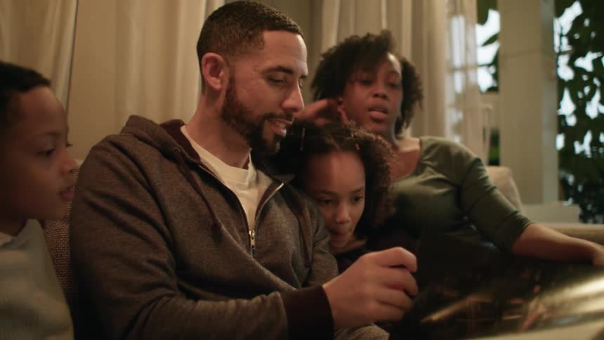 A man and woman sit on the couch with their young son and daughter as the father reads a book to them