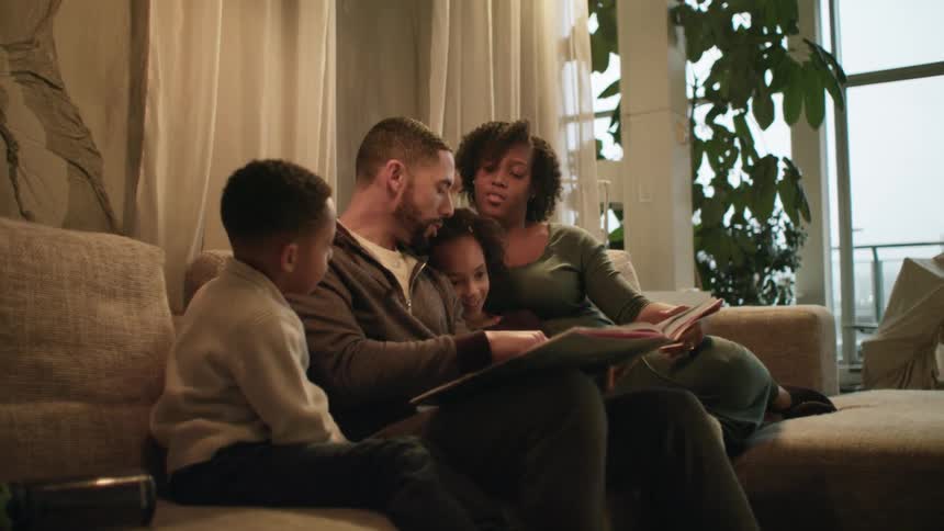 A family sits together on a couch and the father reads to them