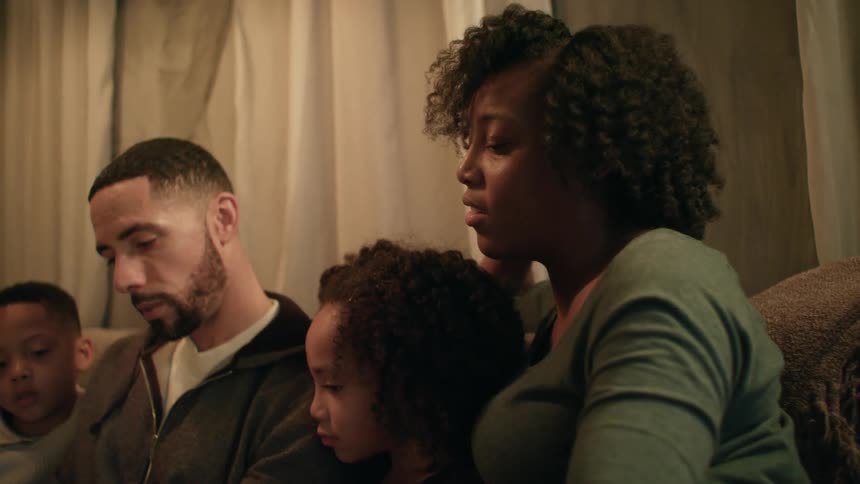 A family sit on the couch while a man reads from a book