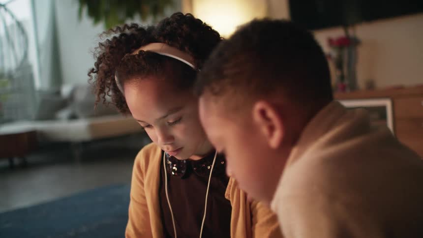 Two young kids sit down on a rug in a living room looking down at a screen