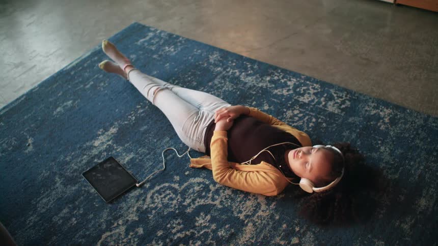 A girl laying on the floor on a carpet listening to music with headphones on a tablet