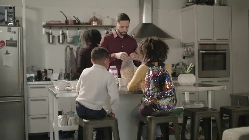 A girl and a boy walking up and sitting on stools in a kitchen with a man and a woman
