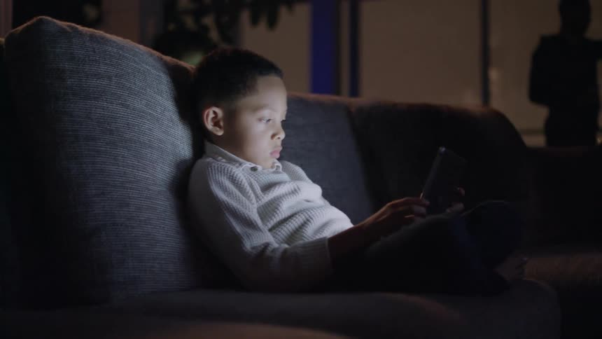 A young boy using a tablet on a sofa in a dark living room