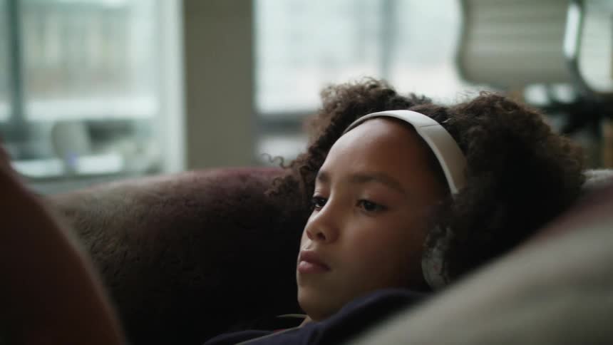 A girl laying on a couch using a tablet with headphones with a window and a fireplace in the background